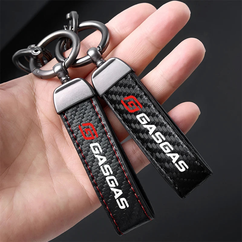 Two carbon fiber keychains with 'GASSGAS' branding held in a hand against a dark background.