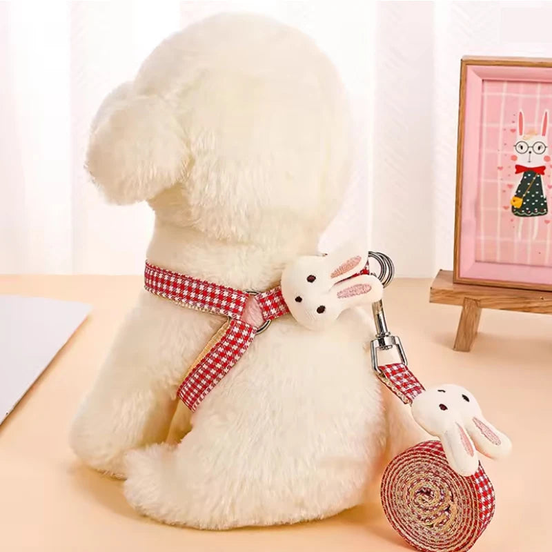 White dog with a red checkered harness and matching leash held by a white rabbit toy, on a light wooden surface.