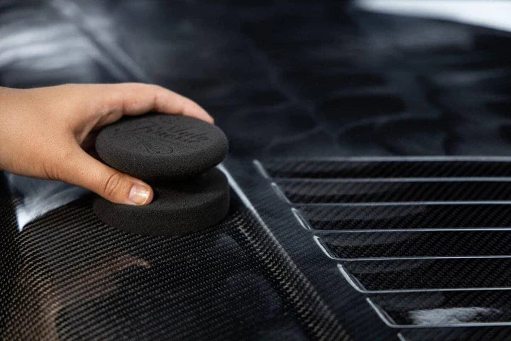Person using a black detailing sponge on a car's carbon fiber surface
