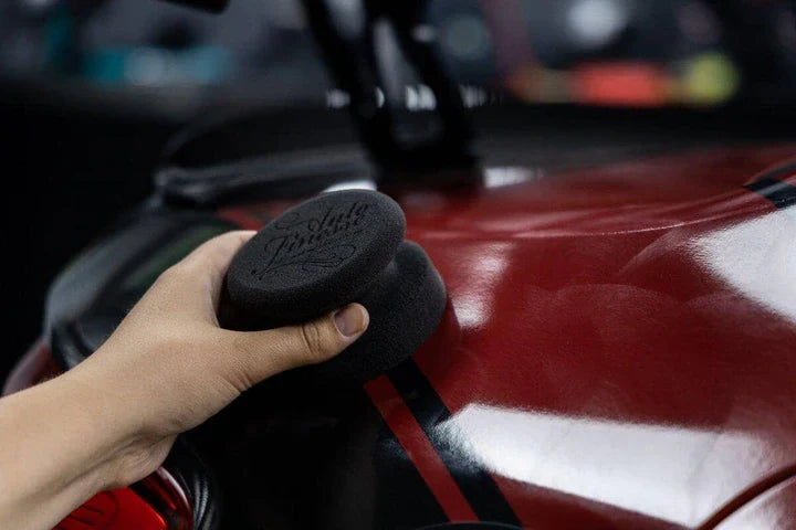 Person holding a black car polish pad with a blurred car exterior background