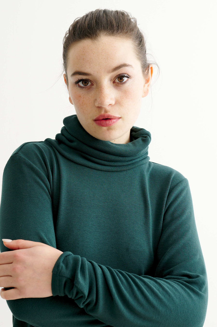 A woman with her hair pulled back wears a dark green long-sleeve turtleneck top against a plain white background.