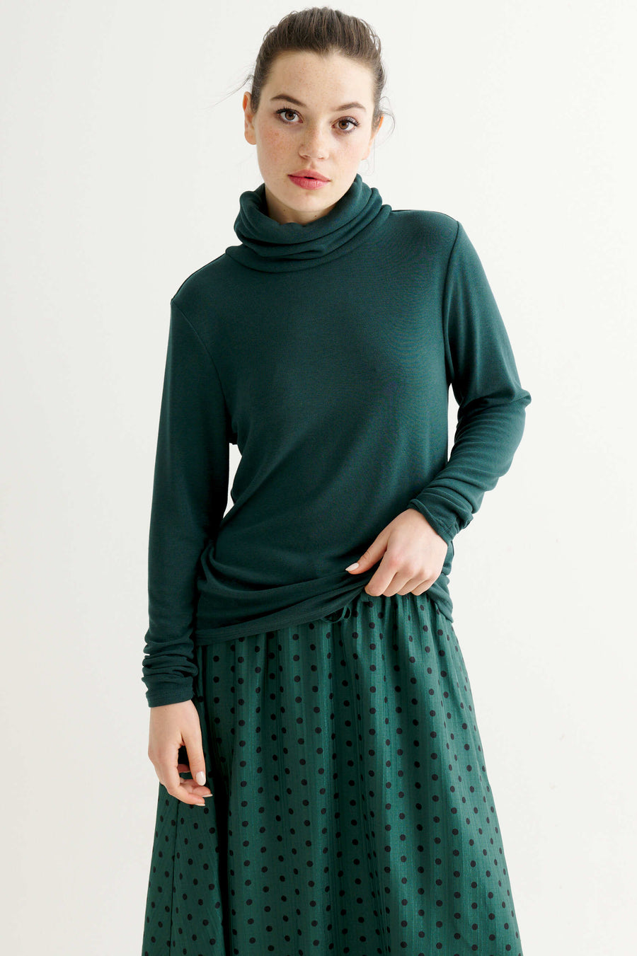 A woman modeling a dark green long-sleeve turtleneck top and a matching dark green skirt with a small polka dot pattern, set against a plain light gray studio background.