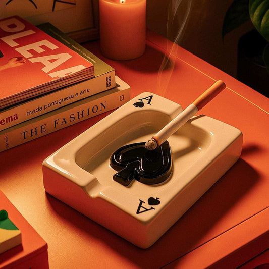 White ceramic ashtray with smoking cigarette on a wooden surface with books and a candle.