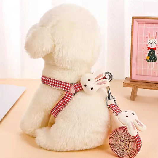 White dog with a red checkered harness and matching leash held by a white rabbit toy, on a light wooden surface.