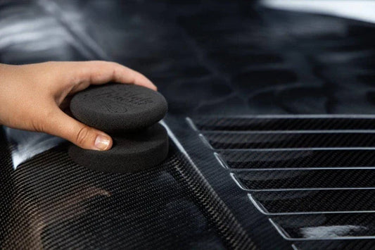 Person using a black detailing sponge on a car's carbon fiber surface