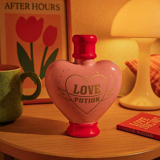 Heart-shaped bottle labeled 'Love Potion' on a table with a mug and book in the background.