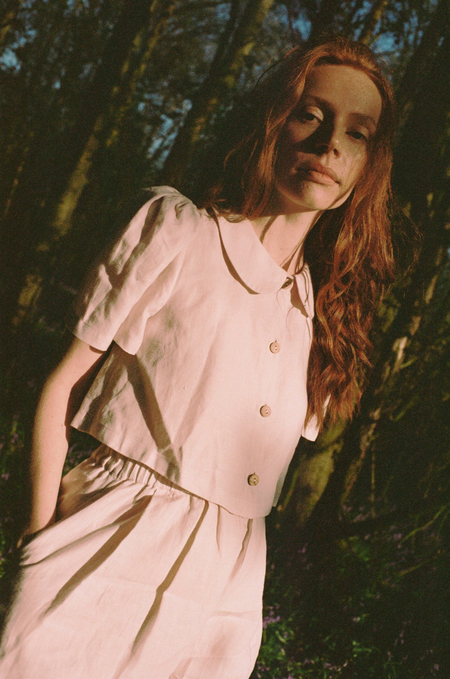 Woman in a light pink blouse and trousers standing among trees