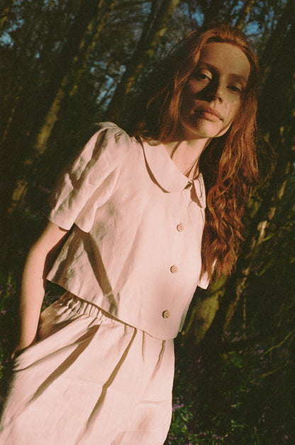Woman in a light pink blouse and trousers standing among trees