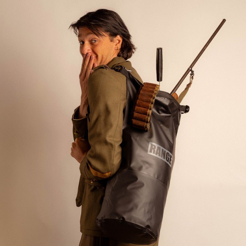Person wearing a green jacket and carrying a 'RANGE' backpack with a rifle and ammunition on a plain background