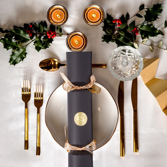 Elegant Christmas table setting with gold cutlery, holly leaves, and candles on a white tablecloth.