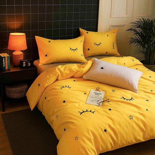 Yellow bedding set with star and eye designs on a bed in a room with a lamp and books.