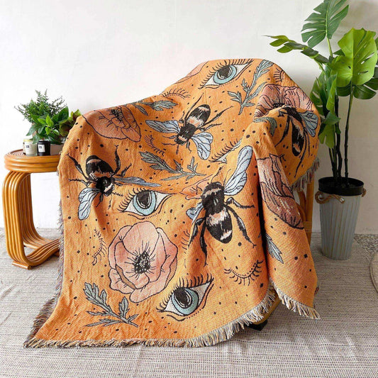 Decorative throw blanket with floral and bee pattern draped over a chair in a room with plants.