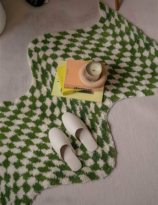 Green and white checkered rug with a stack of books, a small round object, and white slippers on top.